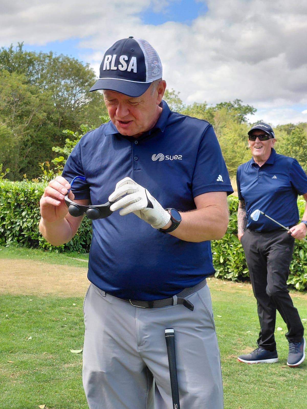 A SUEZ golfer examines a pair of simulation spectacles after wearing them during a golf challenge, while another golfer smiles behind him.