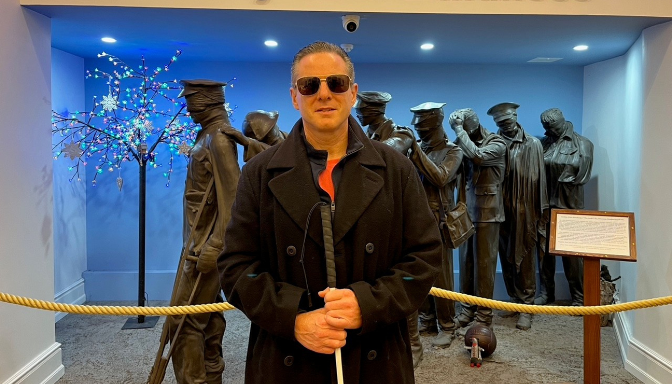 Image of a blind veteran standing before 'The Lads' statue, holding his white cane and wearing dark sunglasses