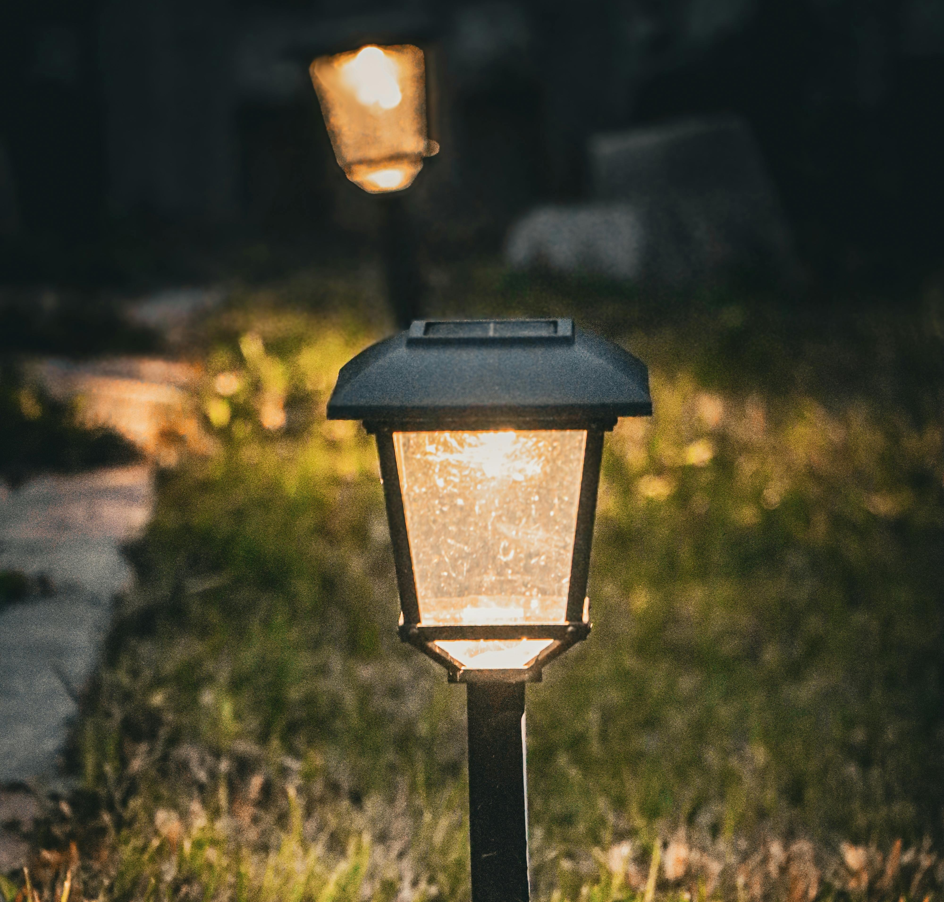 Two outdoor lantern lights illuminate a garden path at dusk, with one lamp in the foreground in sharp focus and another softly glowing in the background among greenery.