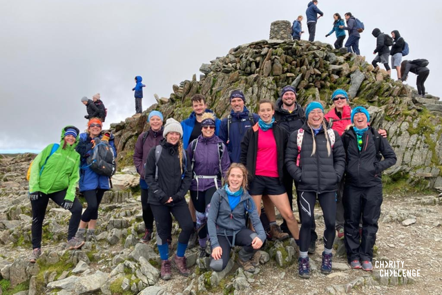 A group of challengers wearing walking gear, smiling widely at the camera.  They are on a plateau of rock just in front of the pile of stones upon which sits the toposcope that marks the peak of Snowdon.