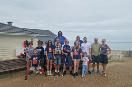 A group photo of Ian with his white cane in the centre surrounded by 13 of his family and friends.