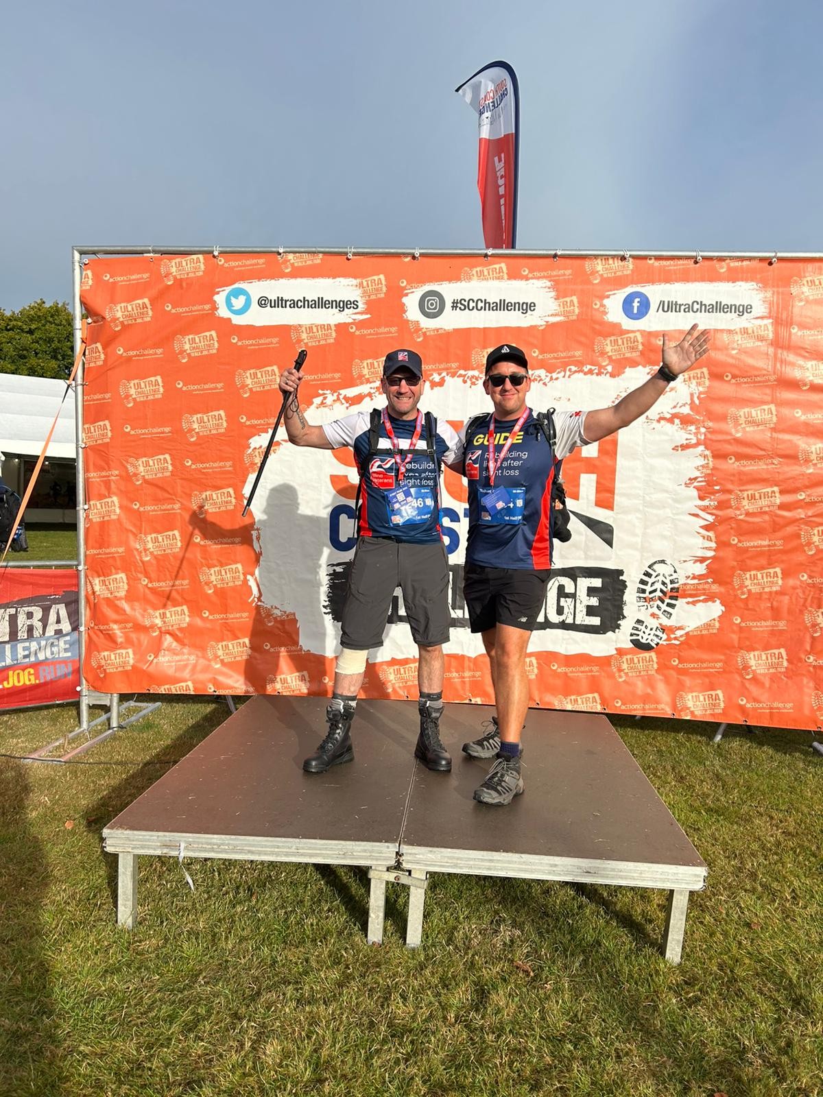 Charlie and Rich stood on a stage with a South Coast Ultra backdrop. The pair are in their Blind Veterans UK t-shirts, shorts and walking boots and have one hand each in the air