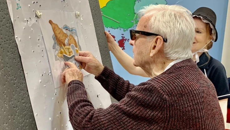 A man wearing glasses stands in front of a board with a picture of fish and chips and a seagull pin.