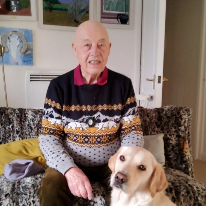Tom is wearing a Christmas jumper and is sitting on the sofa with his guide dog at his feet