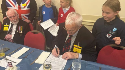 A blind veteran seated and signing autographs for children gathered around him, with a Union Jack in the background.