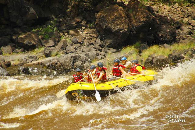A group of people with paddles in a bright yellow inflatable raft in the churning, white-capped waters of the Zambezi river.  The rocky riverbank can be seen in the background.