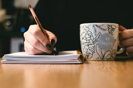 A photo of a person writing on paper while holding white mug with flowers and the words 'be happy' painted on it