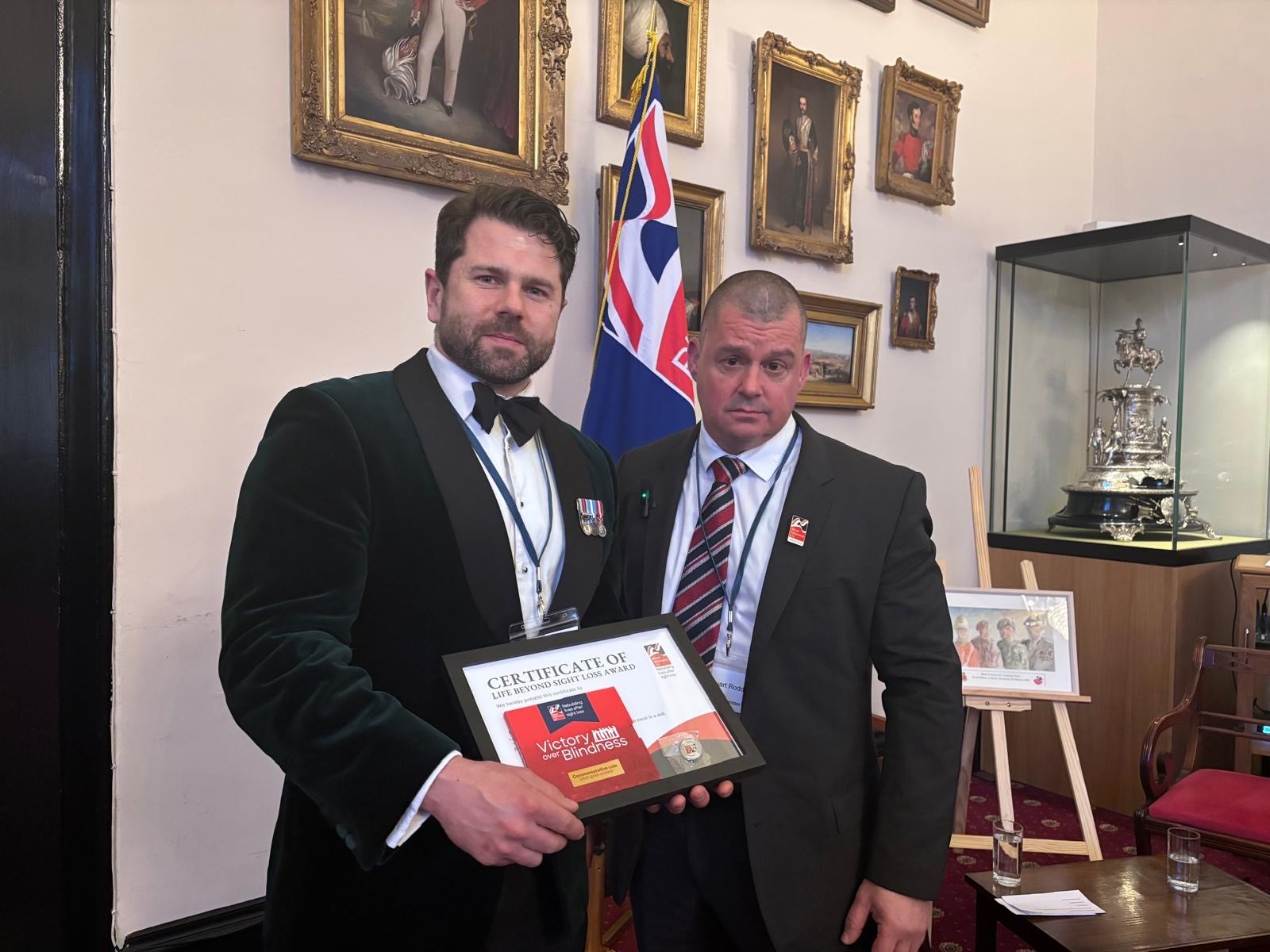Blind veteran Stu and Josh Vause from Amazon stand together in the Indian Army Memorial Room at RMA Sandhurst holding a framed Life Beyond Sight Loss Award certificate, with military portraits and a Union flag behind them.
