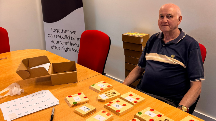 Martin, a volunteer, sits at a table neatly covered in organised craft materials and butterfly house kit components. Behind him is a Blind Veterans UK banner reading “Rebuilding lives after sight loss.”