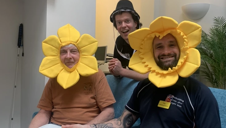Two men sit on a sofa wearing daffodil costumes and a man stands behind them wearing a Welsh bonnet