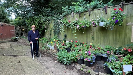 Lawrence stood with a walking stick in one hand a garden fork in the other. His garden shed is behind him and to his left are lots of pots and hanging baskets in bloom with red, yellow, blue, pink and white flowers