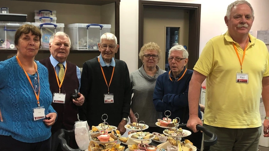 A group of blind veterans smiling, as they stand with a trolley of food they prepared