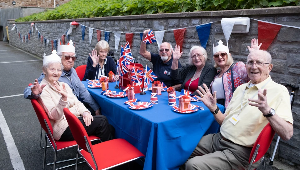 A group of friends sat around a table laden with red, white and blue decorations, waving and smiling for the camera.