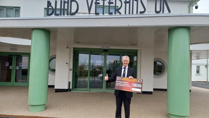 Keven stood outside the entrance of the Rustington Centre of Wellbeing wearing a suit and tie and holding a large cheque