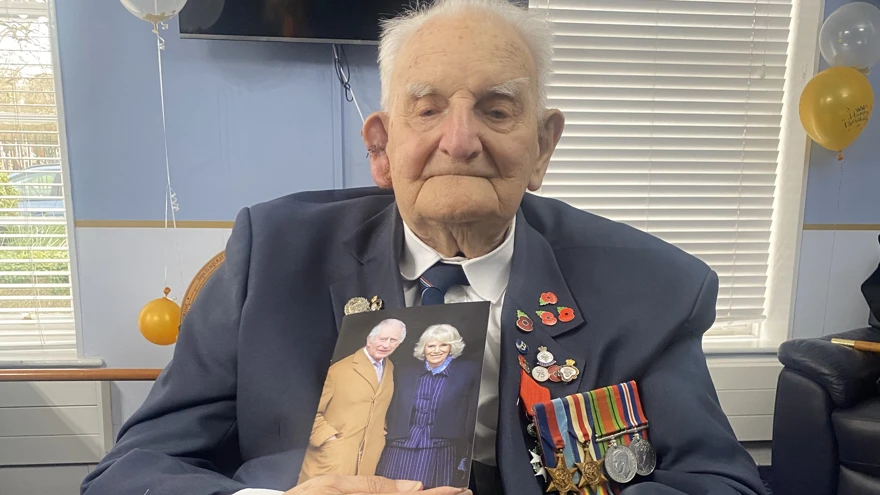 Raymond is wearing his blazer and medals and holds a drink in one hand and his card from the King in the other