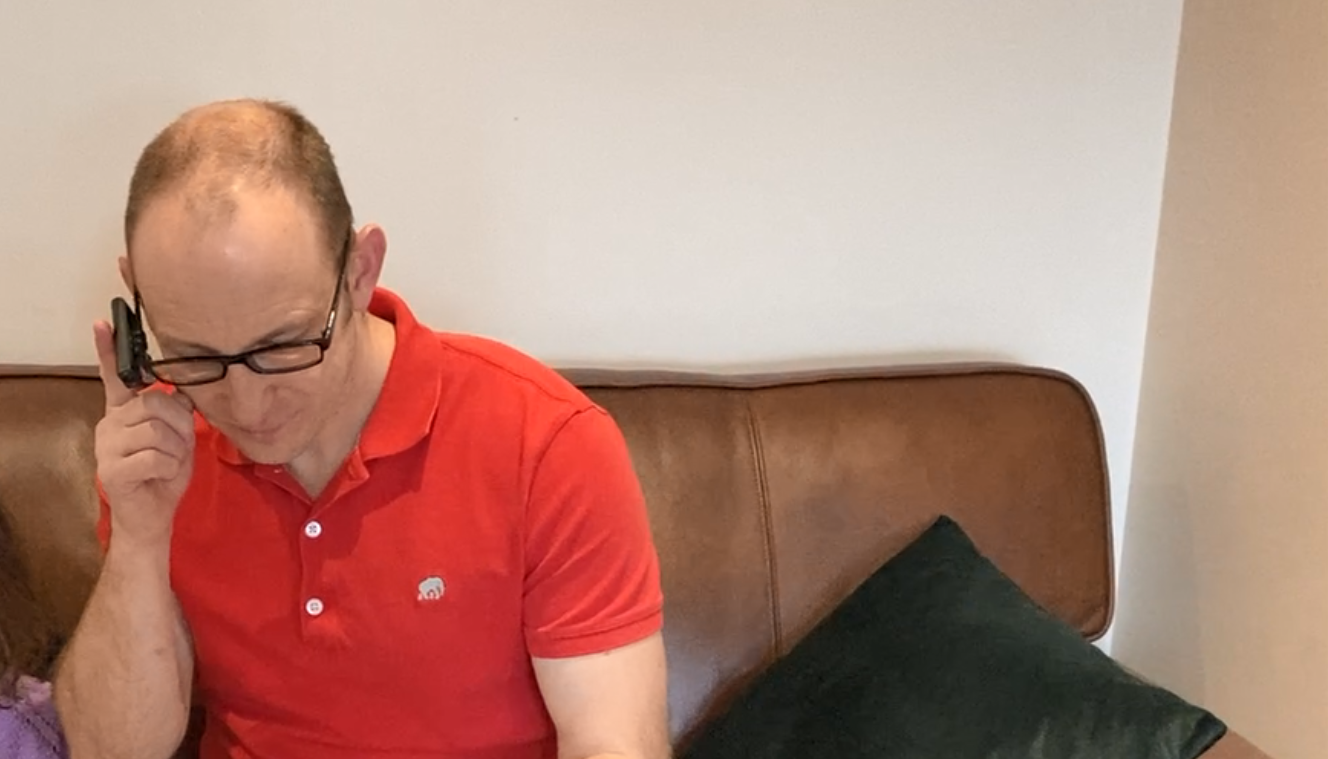 Image shows blind veteran Alan seated, reading a book with the assistance of OrCam glasses