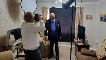 Arthur stands for a photograph in his living room as Richard Cannon holds a large professional camera. In the background, there is a black sheet background and a lighting set up.