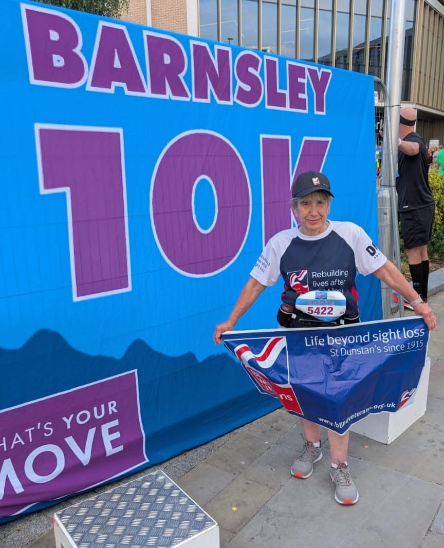 Christine in her Blind Veterans UK Tshirt and baseball cap stood in front of a large Barnsley 10km banner