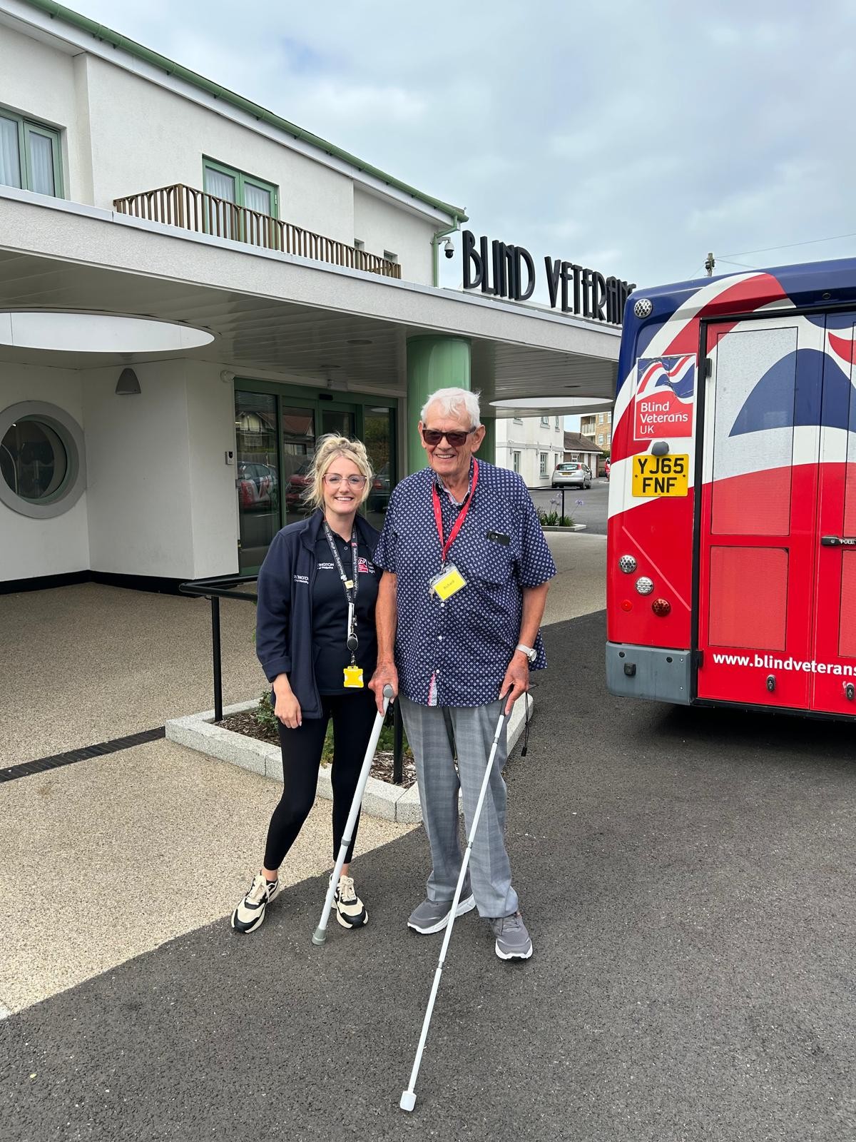 Richard is stood with his white cane outside the front of the Rustington Centre with a Blind Veterans UK bus behind him.
