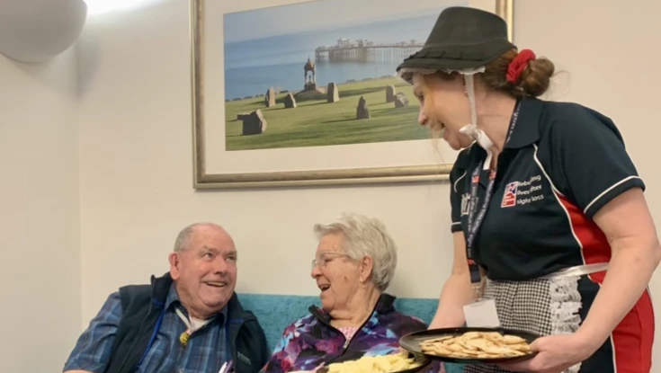 A man and woman sit on a sofa being served cheese by another lady wearing traditional Welsh dress.