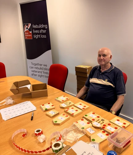 Martin, a volunteer, sits at a table neatly covered in organised craft materials and butterfly house kit components. Behind him is a Blind Veterans UK banner reading “Rebuilding lives after sight loss.”