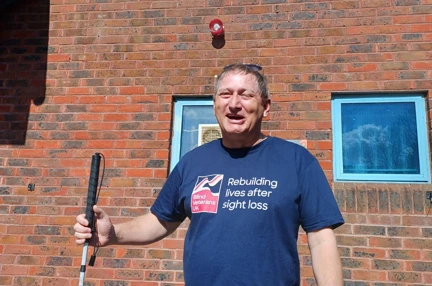 Lee holding his cane and wearing a Blind Veterans UK T-shirt. Lee is smiling outside the swimming pool where he'll be completing his challenge