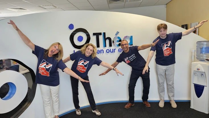 Four members of the Thea team wearing Blind Veterans UK t-shirts are stood in front of a Thea sign with their arms outstretched as if flying