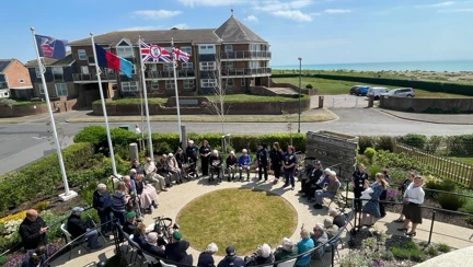 A large group seated in a circle at the Rustington Centre, surrounded by flagpoles and Union Jacks.