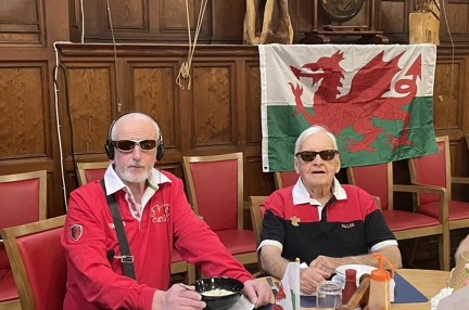 Two men wearing red Wales Rugby shirts sit in front of the Welsh flag which has a white and green background and a red dragon.