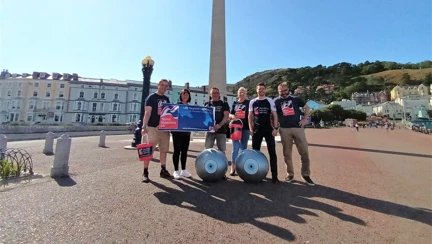 Members of Thea's Surgical Team, wearing Blind Veterans UK branded tops and holding buckets and Blind Veterans UK banners. They are standing in front of the Llandudno Town War Memorial. Two giant yoga balls with eyes painted on them can be seen at the front of the group.
