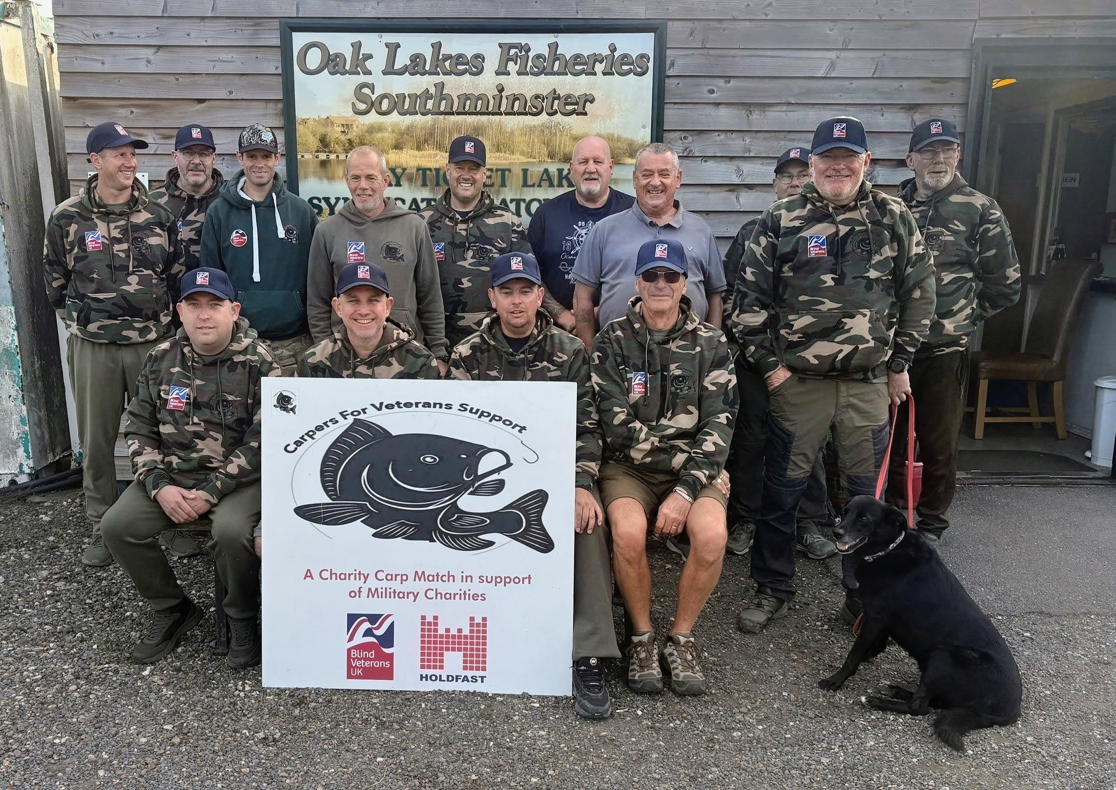 A group photo of the 2026 Carpers for Veterans anglers wearing camouflage tops with Blind Veterans UK logos on them and Blind Veterans UK caps. The group are posed in front of a sign for Oak Lakes Fisheries.
