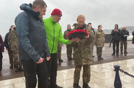 Blind veteran John wearing a bright yellow jacket lays a wreath with a charity volunteer alongside him and a student from the Military Preparation College of training on the other side.