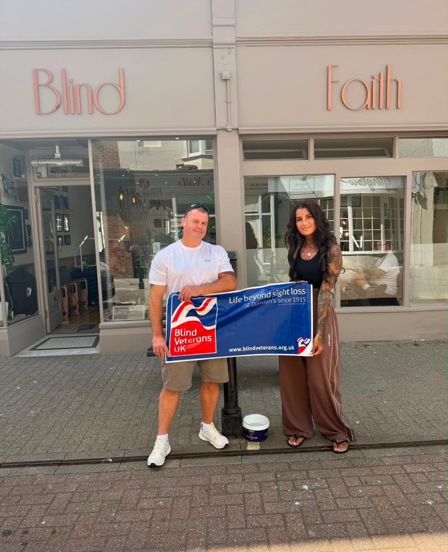Leesa and Stu stood together with a Blind Veterans UK banner outside Blind Faith Tattoos