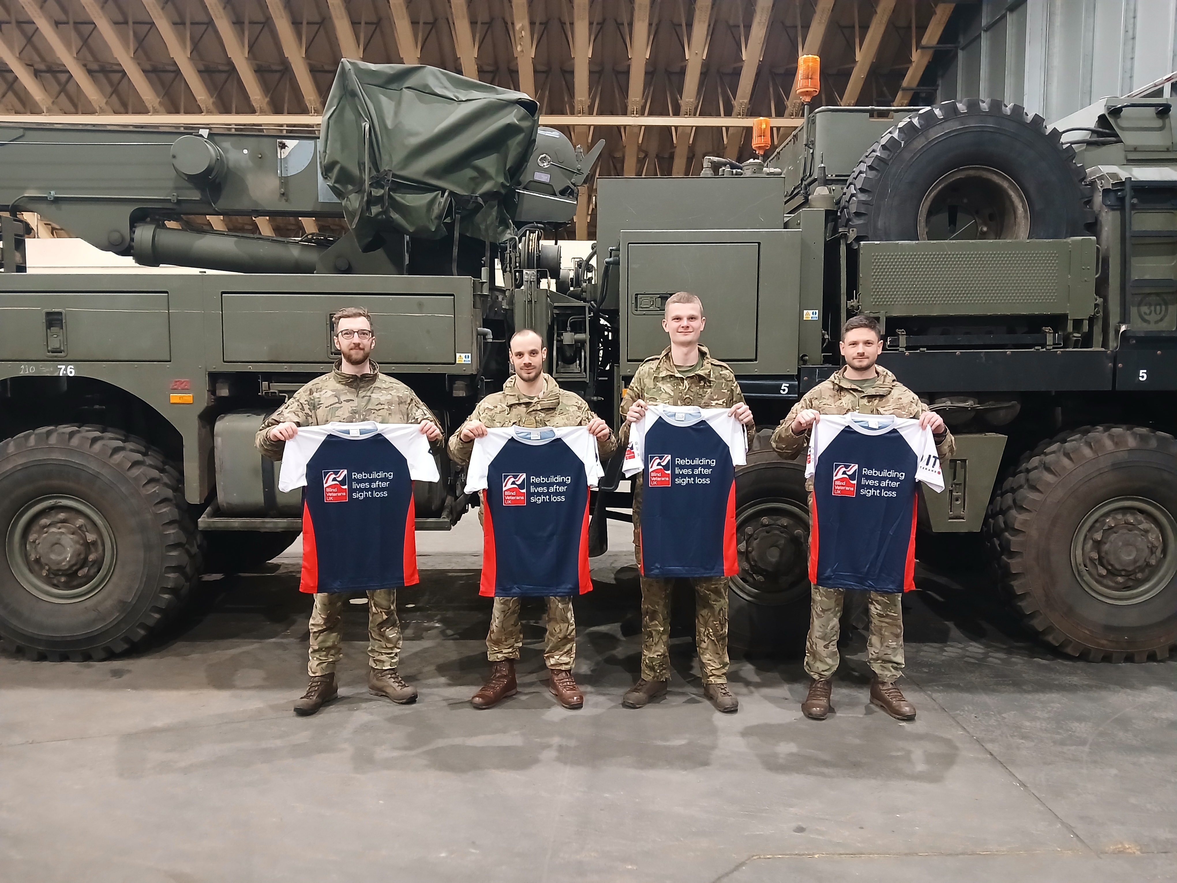 Jack and his Army friends are stood in front of an Army vehicle holding their Blind Veteran UK t-shirts