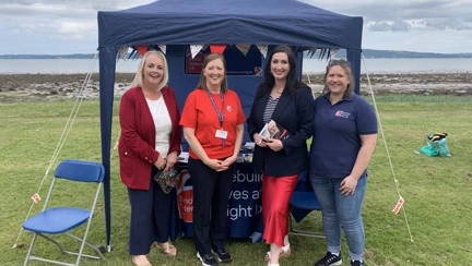 Four women smiling and standing in front of a Blind Veterans UK stall at Armed Forces Day