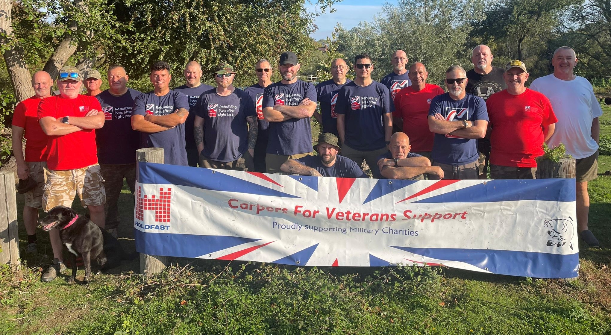 A group photo of all the competitors from this years competition. The group are stood outside on the grass with trees behind them and in front of them is a large banner that reads 'Carpers for veterans support'