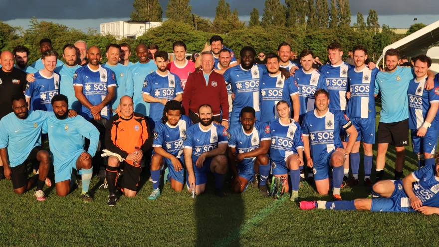 A group shot containing both teams in their football kit and match officials on the pitch. Half the group are kneeling at the front and the others are standing behind with arms around one another's shoulders