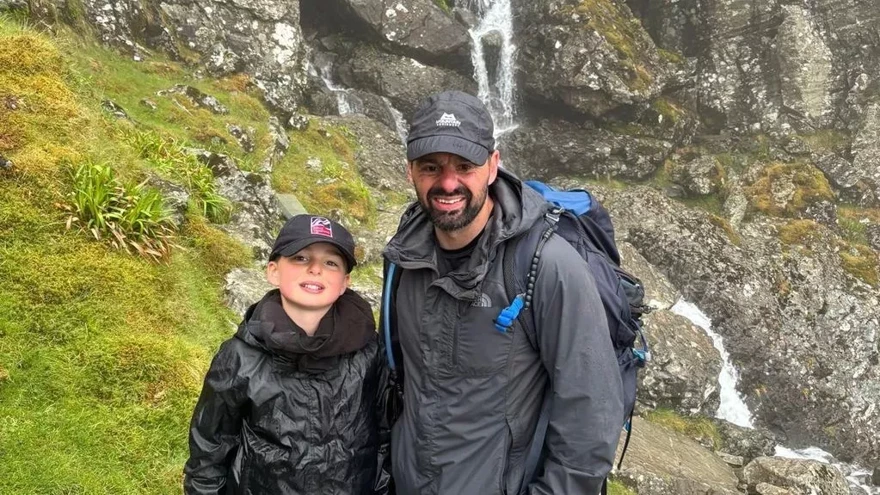 Craig and Max stand out on a mountainside. They are both wearing outdoor gear and baseball caps. They are looking at the camera and smiling.