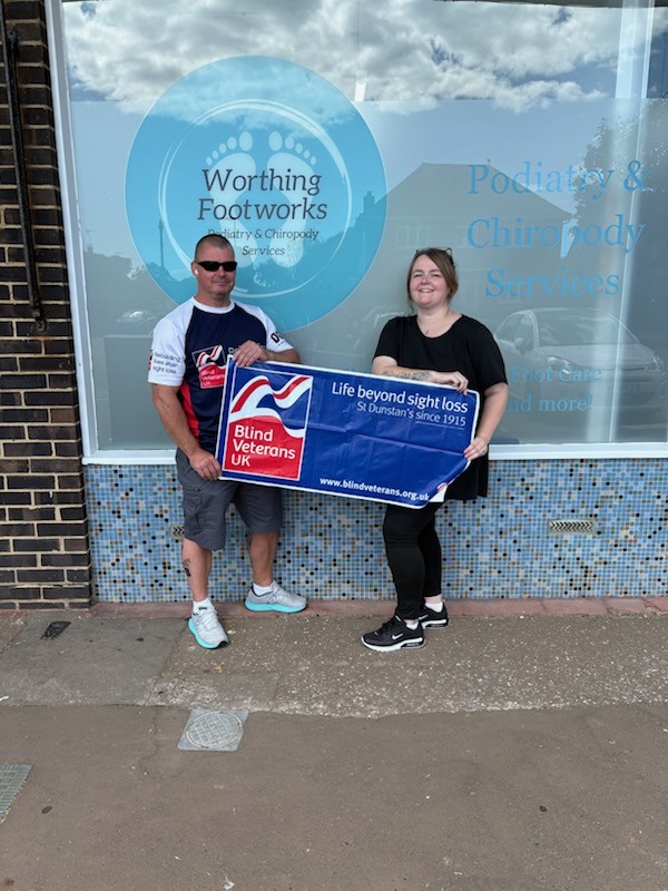 Kate and Stu stand outside Worthing Footworks holding a Blind Veterans UK banner