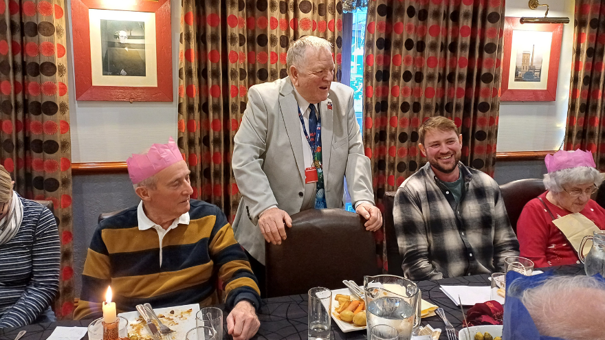 Peter stands to speak at a table in a warmly lit room, blind veterans and staff wearing Christmas hats are seated at the table.