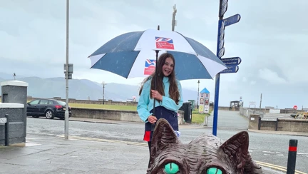 A photo of Emmie, standing next to the Llandudno's Cheshire Cat statue, holding a Blind Veterans UK umbrella