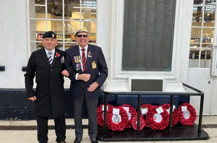 Tony and Danny arm in arm and smiling stood to the side of the war memorial at Brighton Train Station which has eight poppy wreaths laid at its base