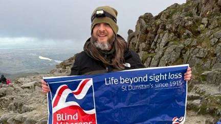 Benjamin at the top of Snowdon holding a Blind Veterans UK banner. Benjamin is wearing a beanie hat and a headtorch.