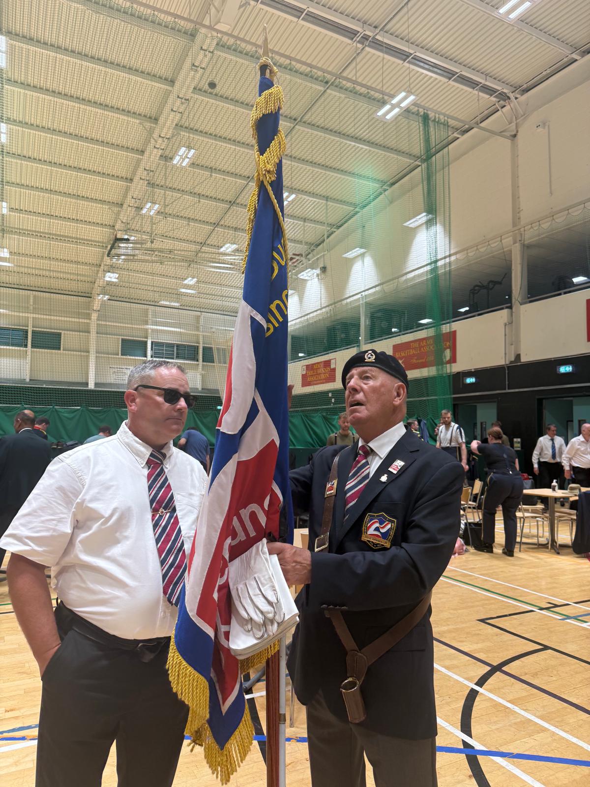 Andy and Brian are stood inside a large sports hall. Andy is in a shirt and tie and Brian is holding the Blind Veterans UK standard