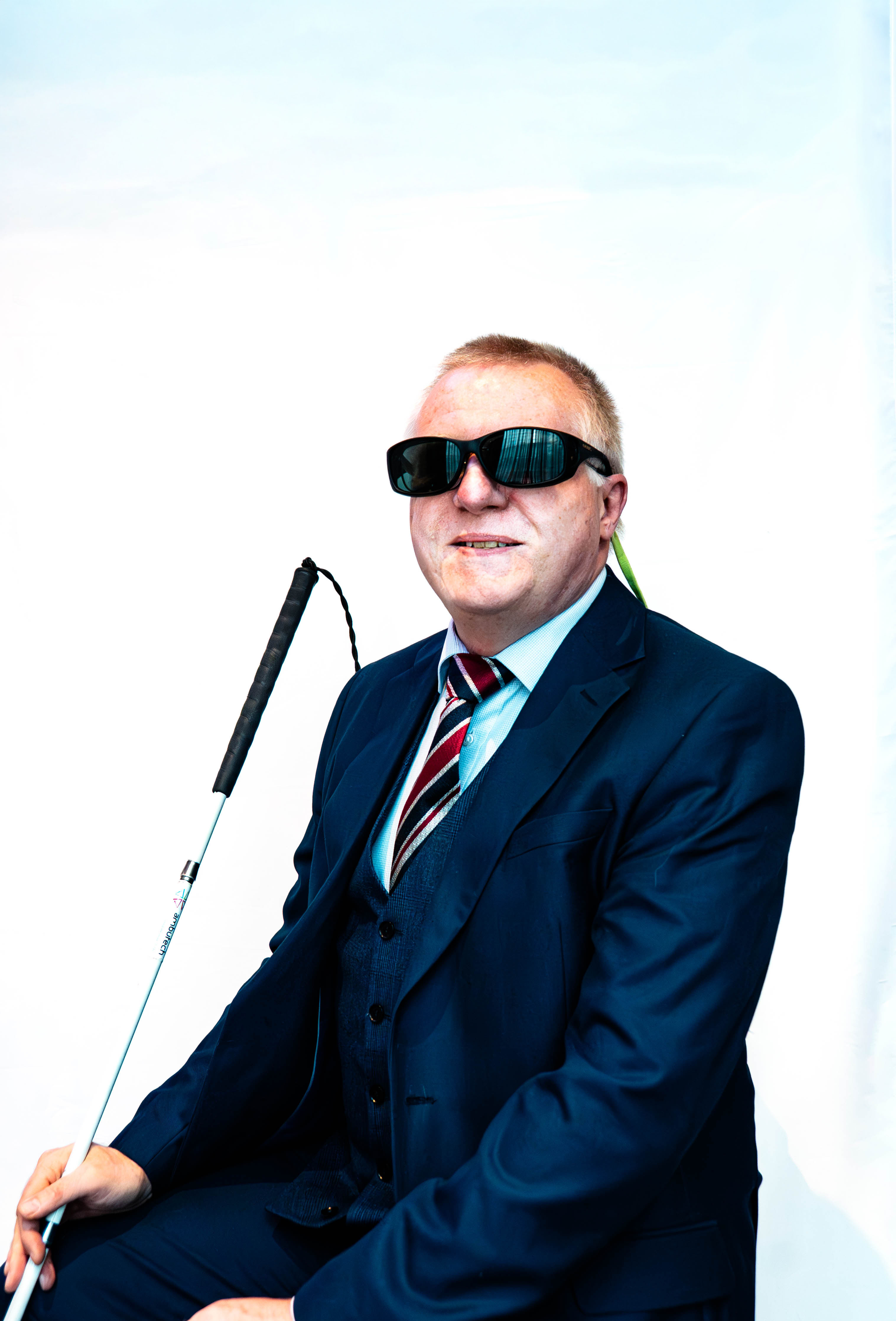 A seated person wearing dark glasses, a navy suit and striped tie holds a white cane against a plain white background.