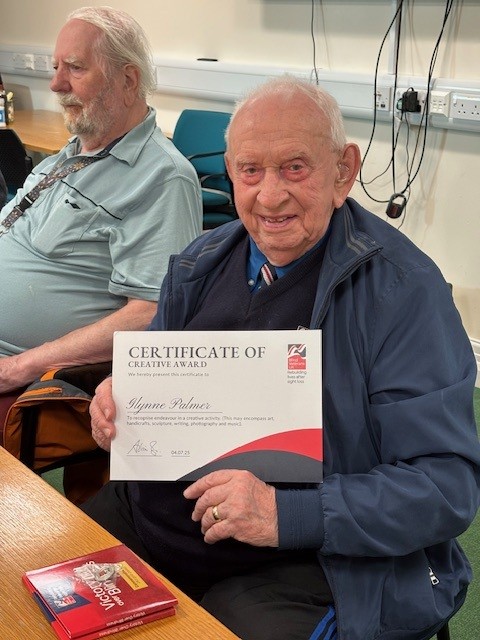 Glynne is seated at a table, smiling as he holds a certificate for creativity.