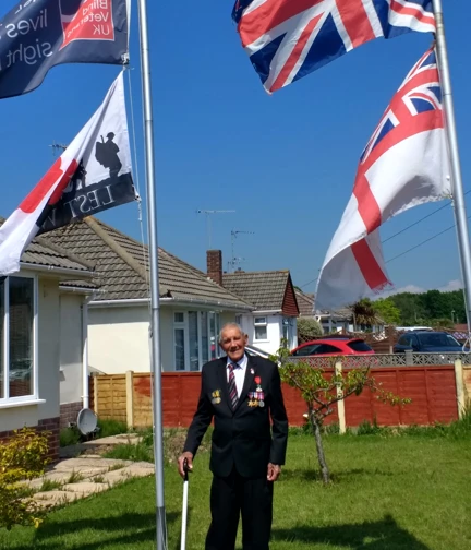 Blind veteran Eddie standing between two flag poles in his garden, displaying a Blind Veterans UK, Union Jack and Lest We Forget flag