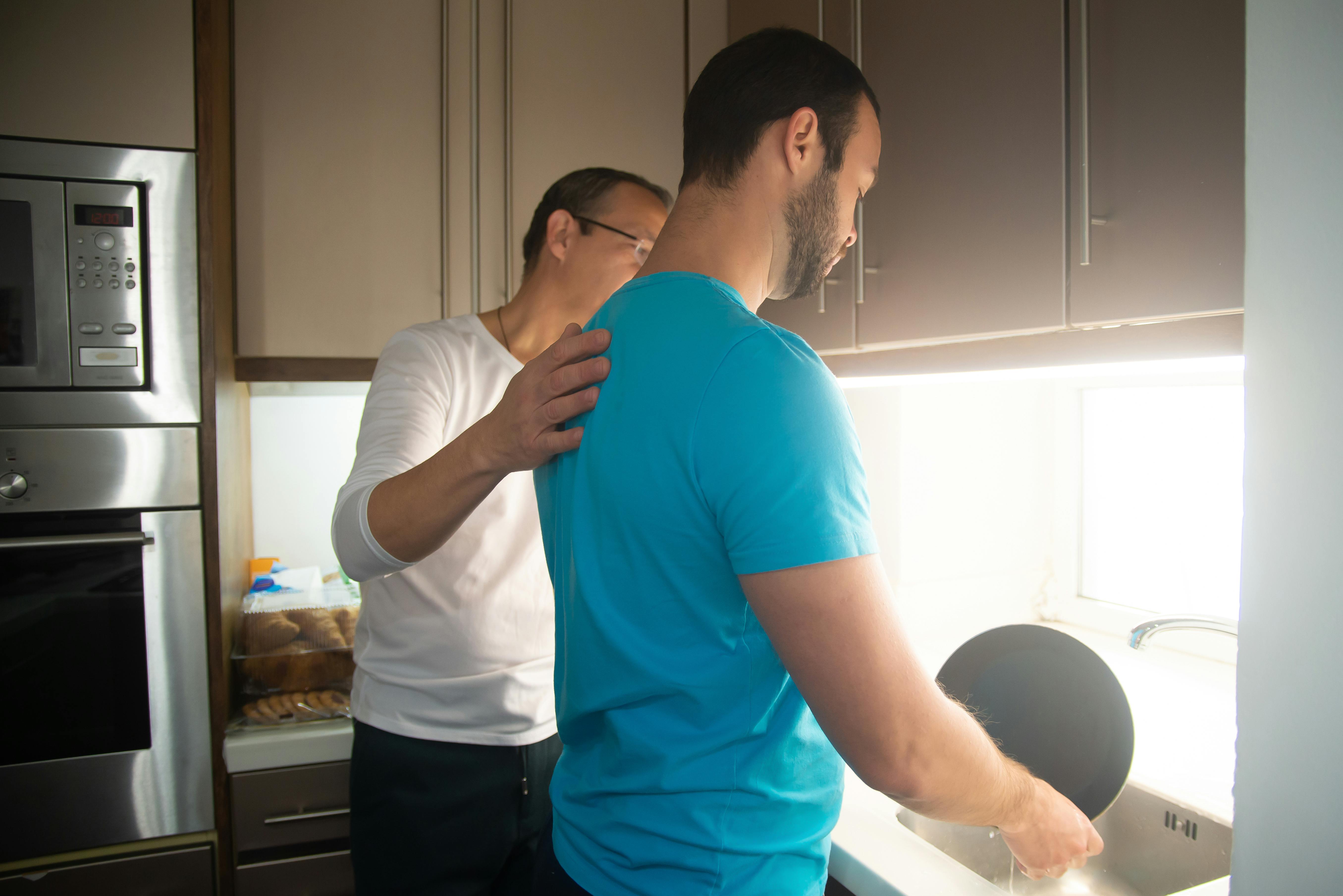 Two people standing in a kitchen at a sink, washing dishes; one person gently places a hand on the other person’s shoulder for guidance, with cupboards and a built-in oven visible in the background.