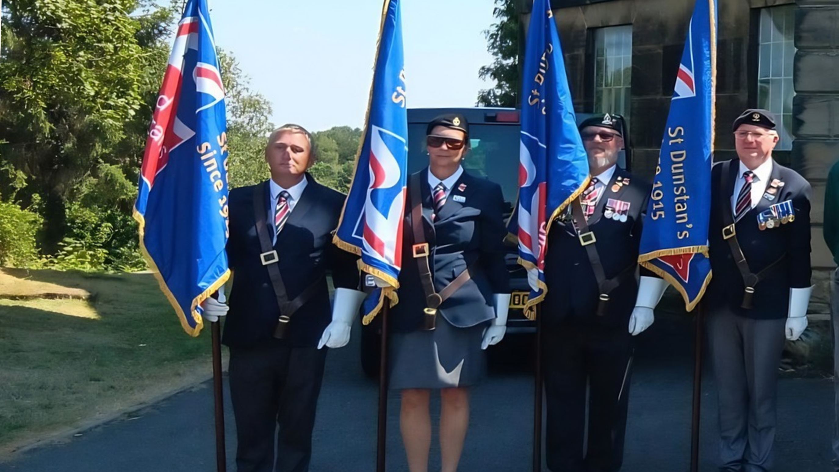 Four standard bearers stand outdoors in formal uniforms, berets and white gloves, each holding a blue standard with gold edging. The person second from the left is positioned prominently between two standards.