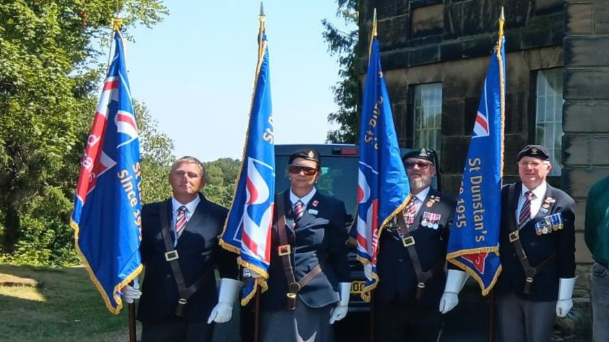 Four standard bearers stand outdoors in formal uniforms, berets and white gloves, each holding a blue standard with gold edging. The person second from the left is positioned prominently between two standards.